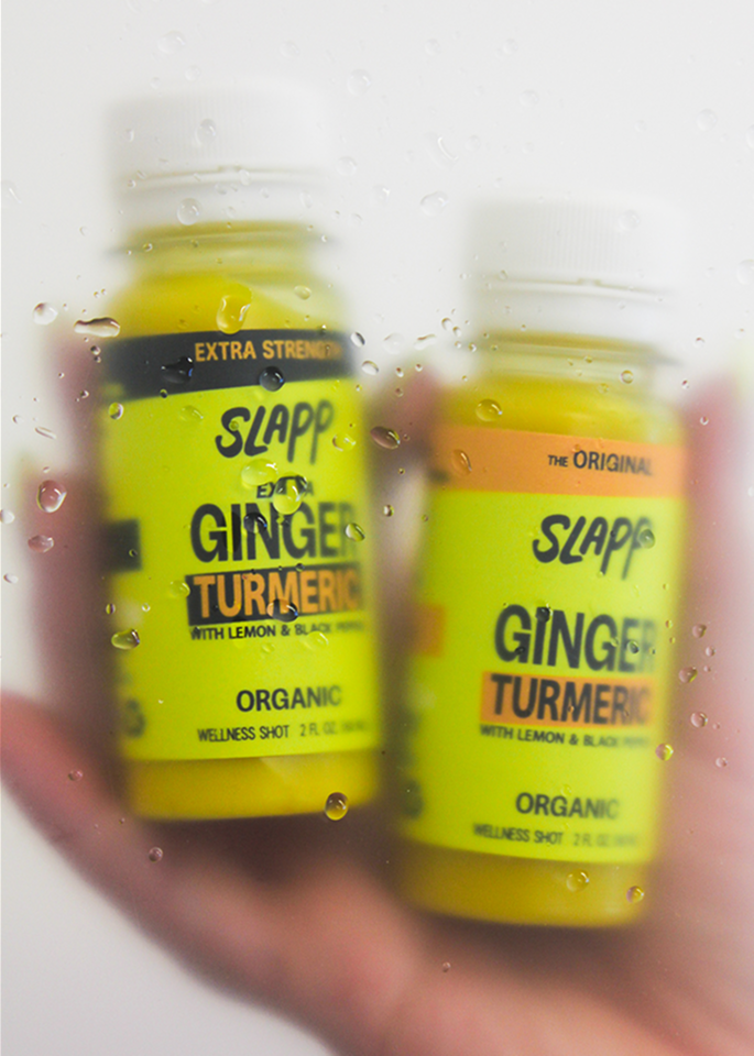 Two Slapp Ginger Turmeric wellness shot bottles held in hand behind a wet glass surface.